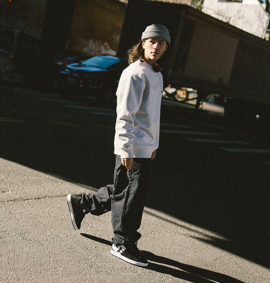 A person wearing a gray beanie, white sweatshirt with a subtle DC Shoes logo, dark pants, and DC Shoes Men’s Court Graffik Shoes walks on a sunlit street, casting a puffy silhouette in their shadow.
