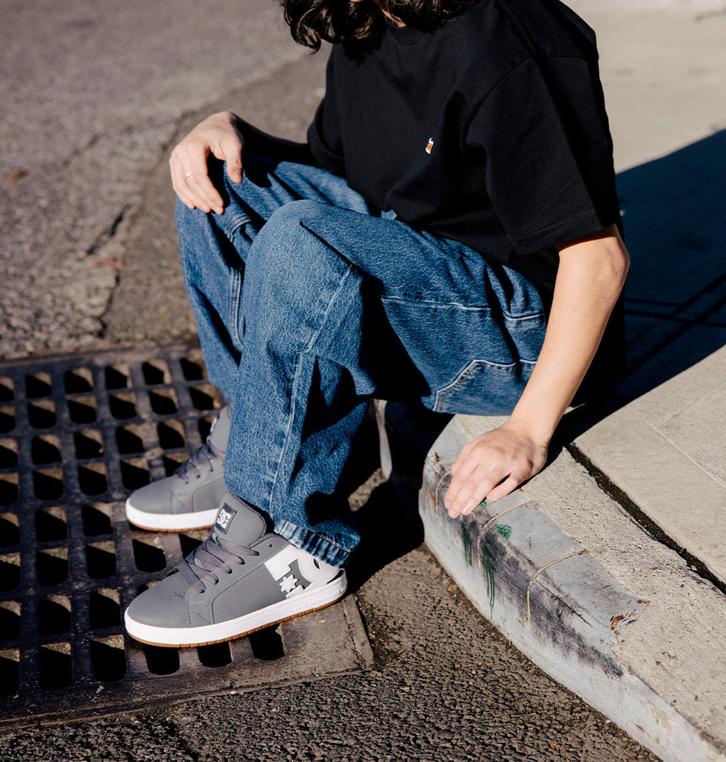 A person in a black t-shirt and blue jeans sits on a curb with one hand resting on the sidewalk and the other on their knee. They are wearing gray DC Shoes Men’s Court Graffik shoes featuring chunky styling with white and black accents.