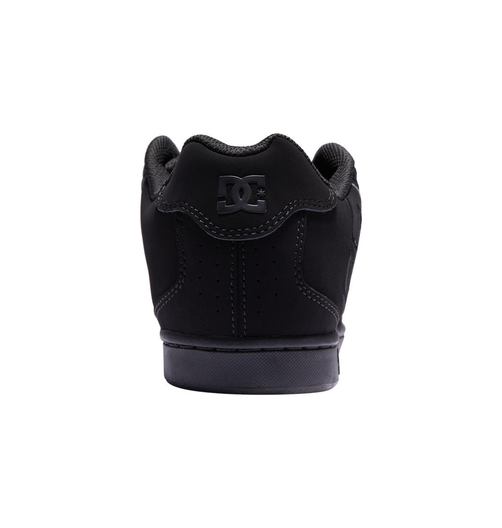 Back view of a black DC Shoes Men’s Net Shoe with the DC logo displayed on the heel. This puffy sneaker showcases a padded collar, textured midsole, and perforations for ventilation. Made from durable fabrics, it is set against a plain white background.