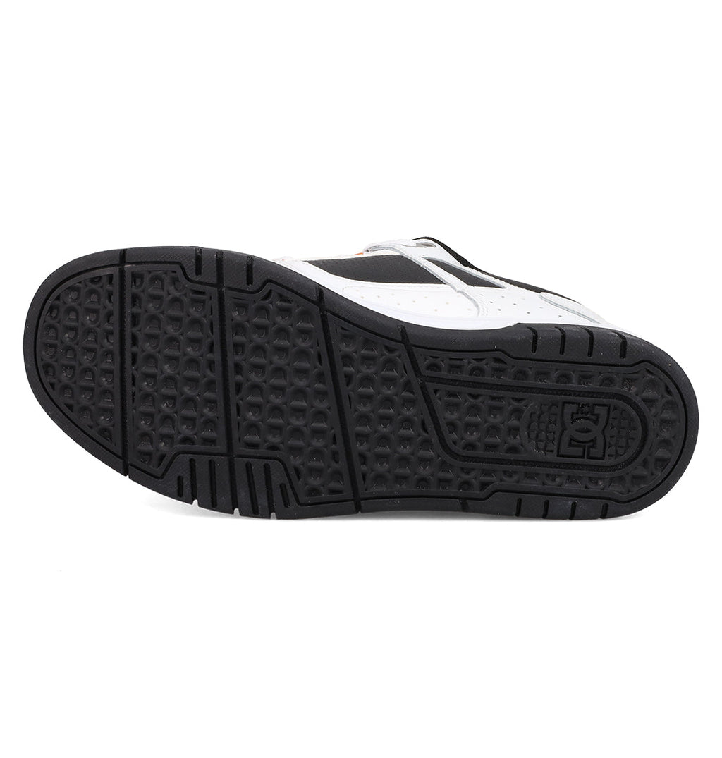 The image shows the black rubber outsole of DC Shoes Men's Stag Shoes, with geometric tread patterns, a grid-like texture, and a circular logo by the heel. The durable cupsole pairs with a white and black upper inspired by skate culture.