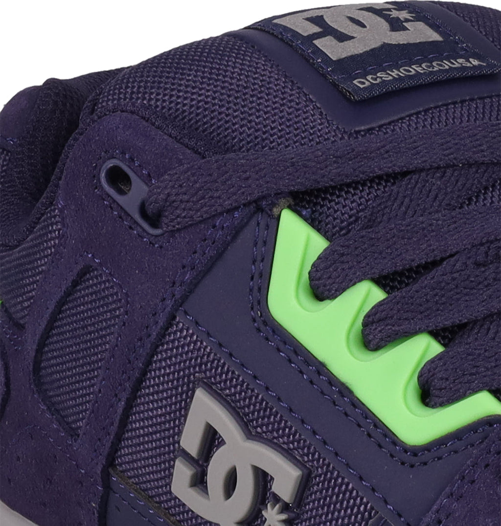 Close-up of DC Shoes Men's Stag skate shoe in purple with a cupsole, textured upper, gray DC logo, dark laces, and bright green accents. The tongue features a visible "DC SHOES" label for classic skate style.