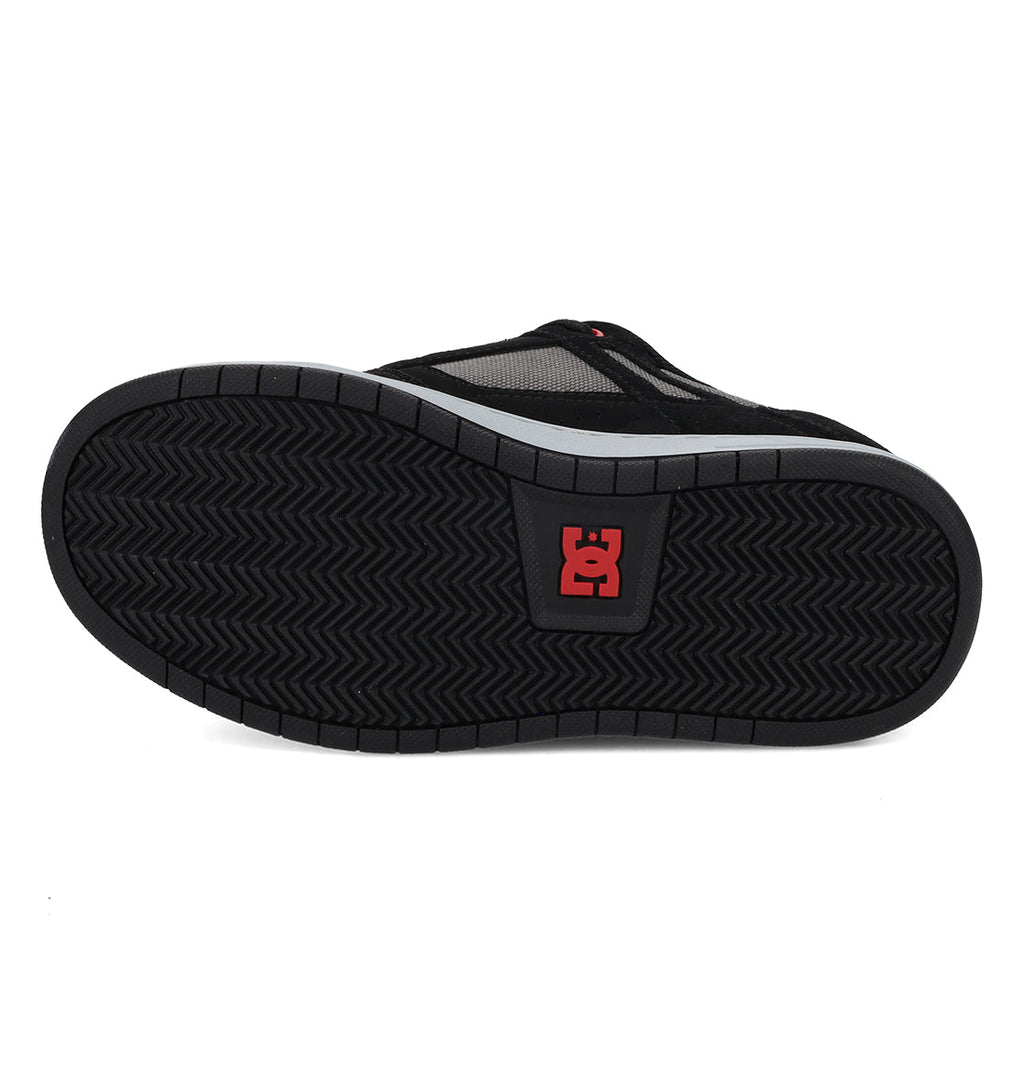 Bottom view of the DC Shoes Kids’ Stag Shoe in black, showcasing a herringbone tread pattern and a red DC Shoes logo near the center of the durable rubber sole, designed for enhanced impact absorption and lightweight cushioning.