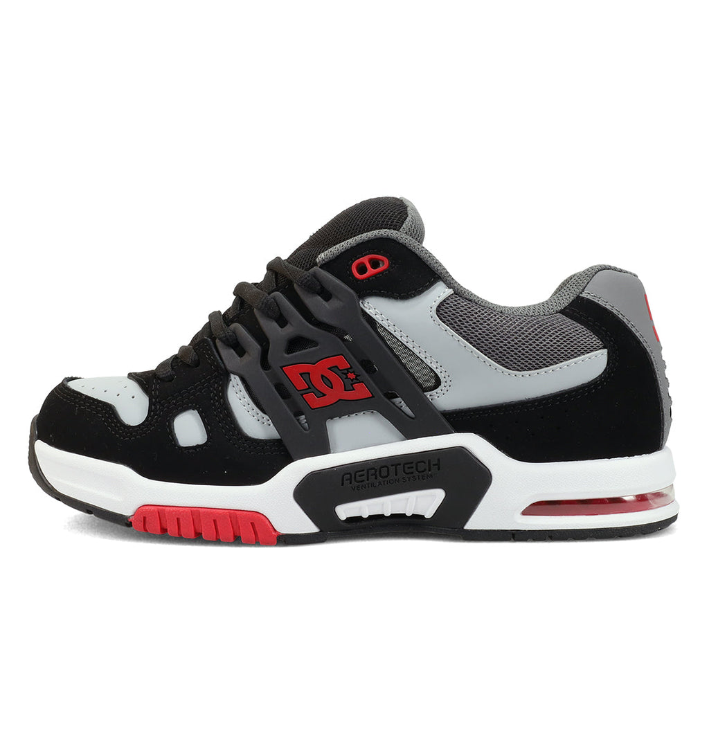 The DC Shoes Men’s AT-2 Shoes feature a black, grey, and red colorway with mesh panels for breathability and a thick sole. Designed for comfort and performance, these skate shoes showcase the DC logo on the side, bold red detailing near the toe and heel, and an AEROTECH ventilation system. The sole is finished with ACROTECH text, giving them a distinctive look.