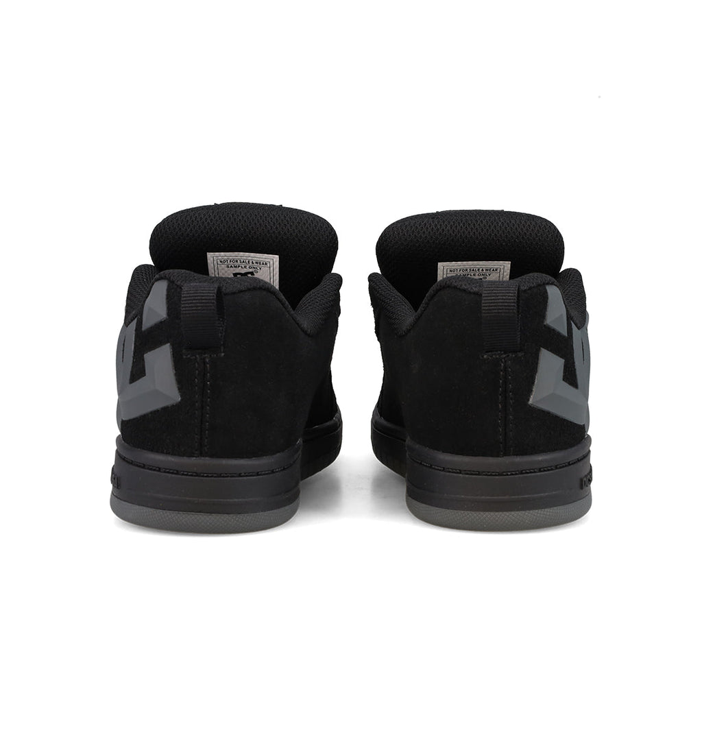 A pair of Kids’ Court Graffik Beveled Special Edition Shoes by DC Shoes are shown from the back, featuring thick black soles and a dark geometric beveled TPR rubber logo on each heel for signature DC branding. The black low-top sneakers are positioned side by side against a white background.