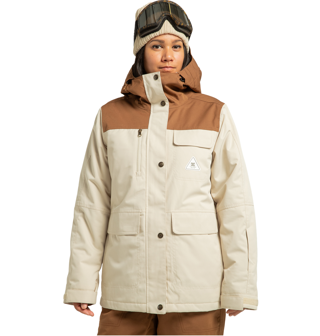 A person stands against a plain white background ready for any winter adventure, dressed in the DC Shoes Women’s Liberate Insulated Jacket—crafted from recycled polyester in beige and brown—paired with brown pants, a beanie, and ski goggles on their head.