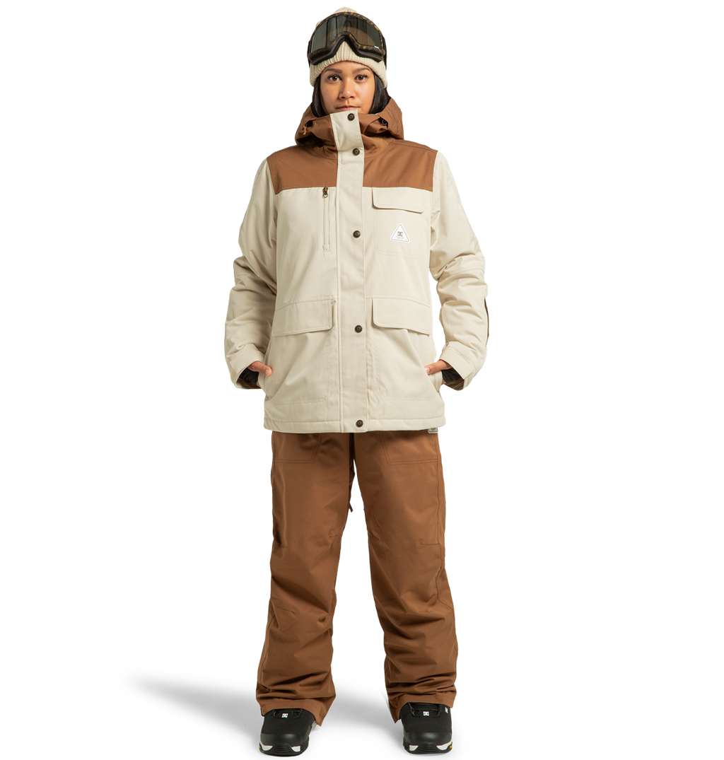 A person stands against a plain white background, ready for a winter adventure in the DC Shoes Women’s Liberate Insulated Jacket and matching beige and brown pants made from recycled polyester, complemented by black gloves, black boots, and ski goggles resting on their head.