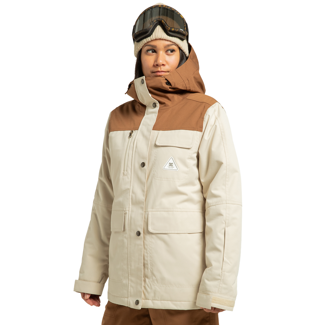 A person models the DC Shoes Women’s Liberate Insulated Jacket in beige and brown, crafted from recycled polyester, paired with brown pants and ski goggles atop a knit hat, gazing to the side against a plain white background—ready for any winter adventure.