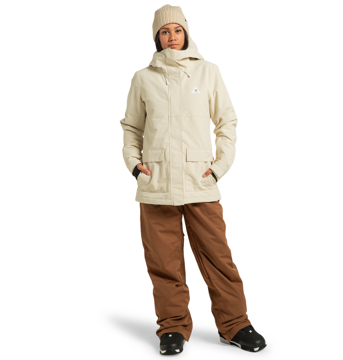 A woman stands with her hands in the pockets of her DC Shoes Women’s Cruiser Insulated Jacket, crafted from recycled polyester and featuring advanced weather defense technology, paired with brown pants, black boots, and a beige beanie against a plain white background.