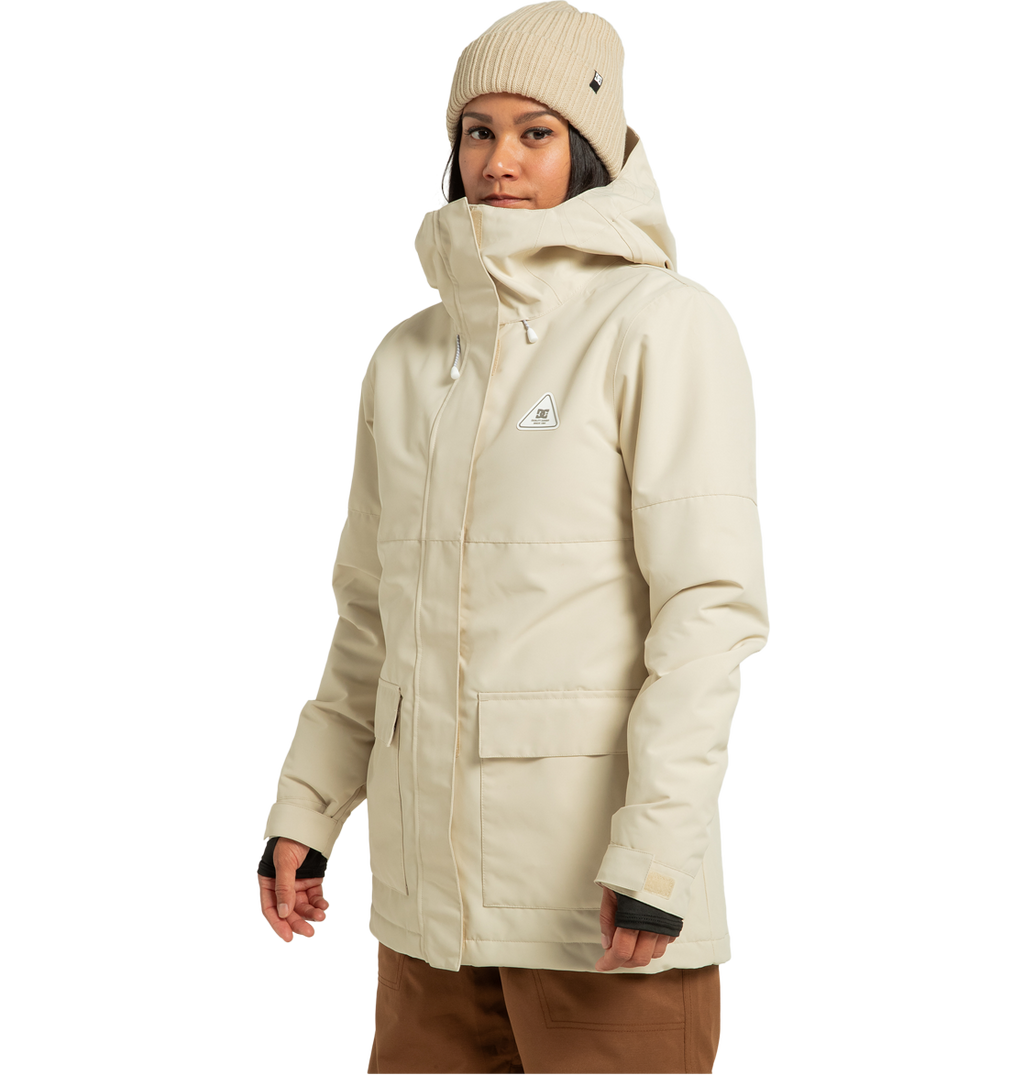 A woman stands against a plain white background, facing slightly to the side with a neutral expression, wearing the DC Shoes Women’s Cruiser Insulated Jacket in beige—crafted from recycled polyester and featuring weather defense technology—paired with brown pants and a beige knit beanie.