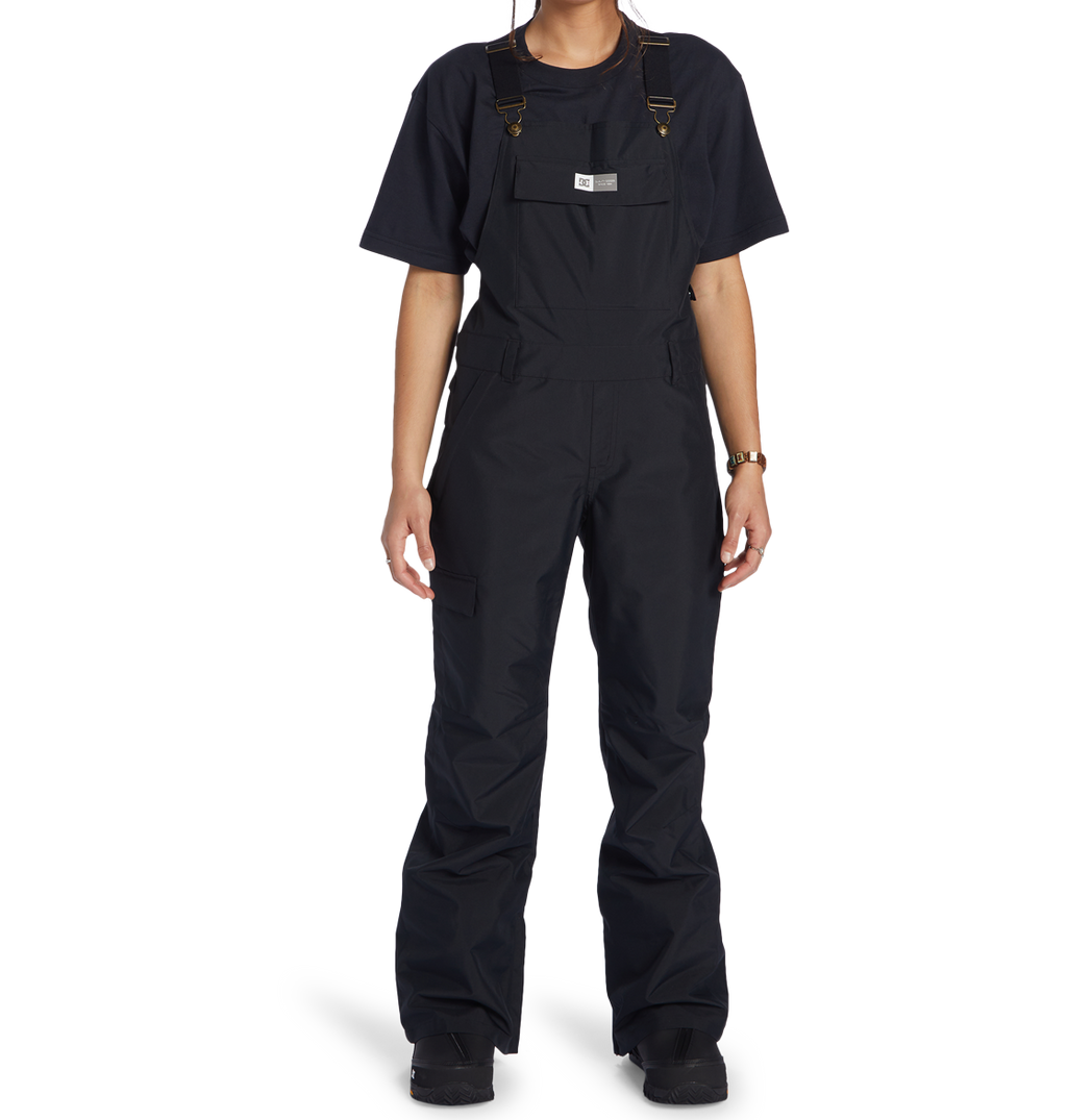 A person, their head partially out of frame, models the DC Shoes Women’s Valiant Bib—black overalls made from recycled polyester—layered over a black t-shirt, set against a plain white background.