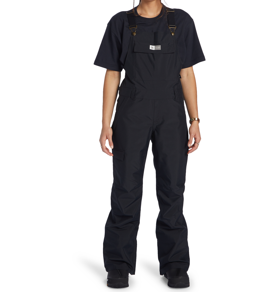 A person, their head partially out of frame, models the DC Shoes Women’s Valiant Bib—black overalls made from recycled polyester—layered over a black t-shirt, set against a plain white background.