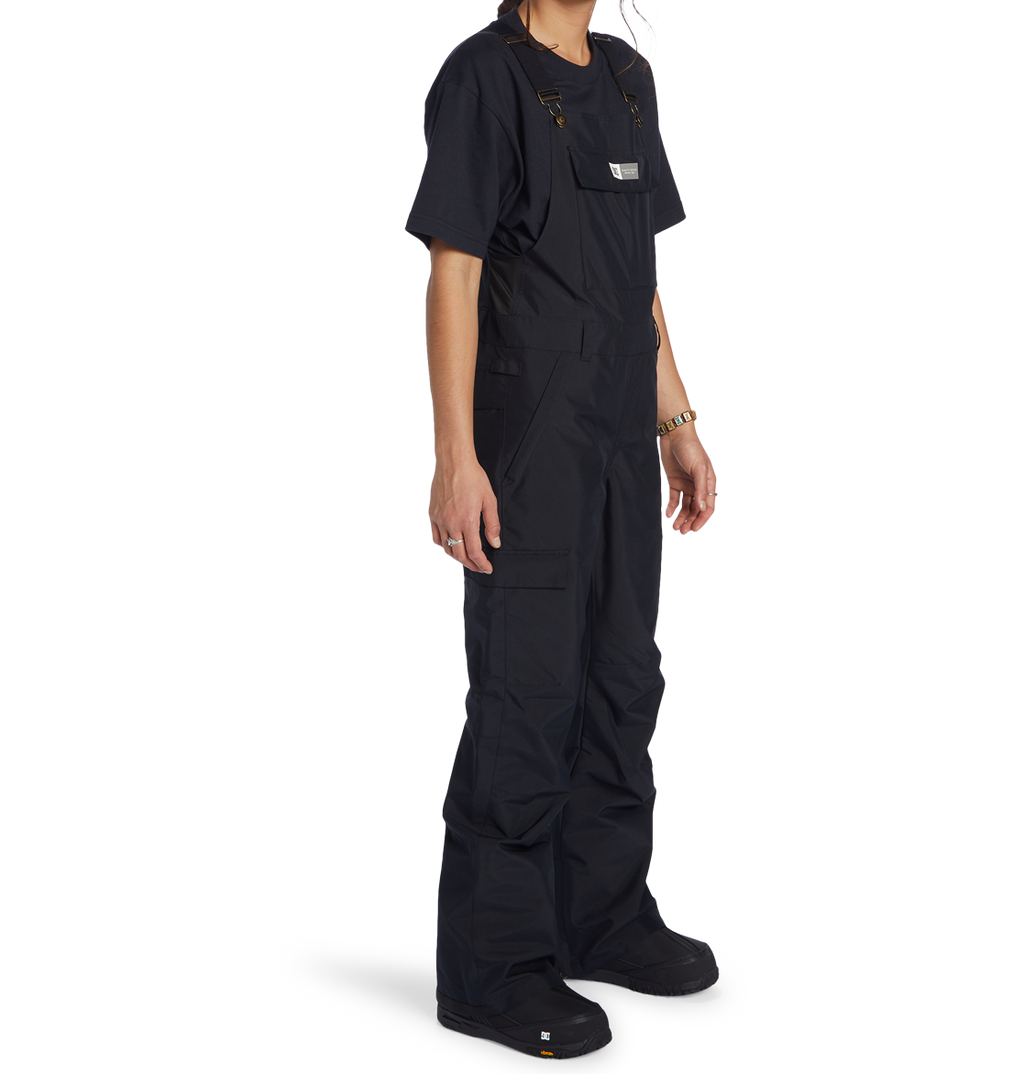 A person stands facing slightly right against a plain white background, wearing a black T-shirt and the DC Shoes Women’s Valiant Bib with pockets, crafted from recycled polyester. Their face is not visible.