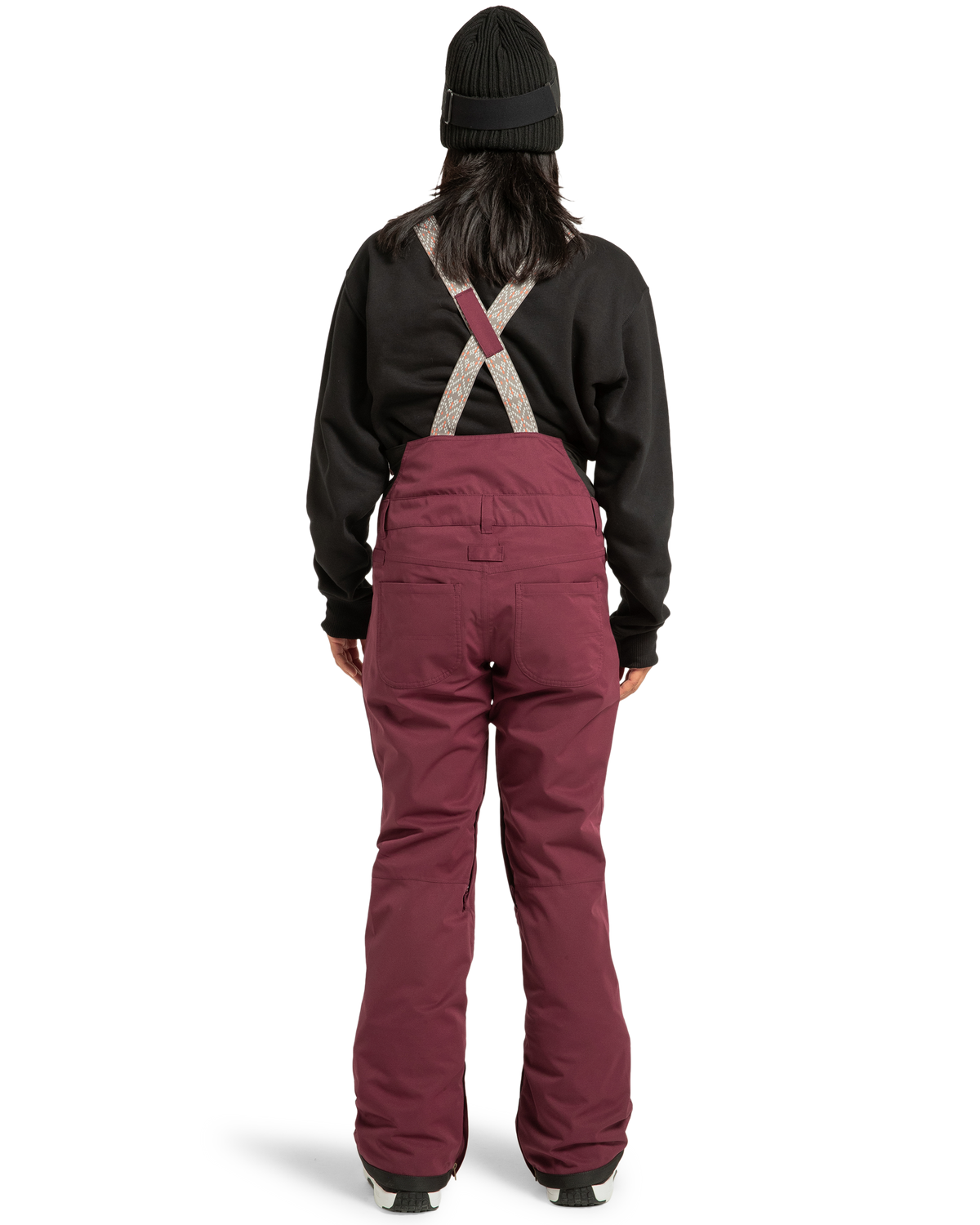 A person stands with their back to the camera, wearing a black beanie, black sweatshirt, and maroon DC Shoes Women’s Crusade Shell Bib snow pants featuring suspenders. Set against a plain white background, this stylish and eco-friendly bib is perfect for women seeking sustainable ski adventures.