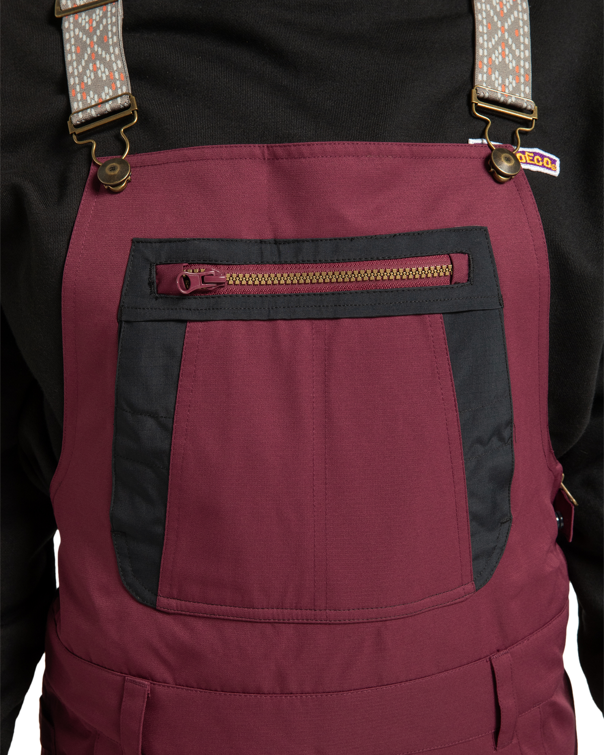 Close-up of a person wearing the DC Shoes Women’s Crusade Shell Bib in maroon, featuring a zippered front pocket and patterned adjustable straps layered over a black top. Only the torso area is visible.