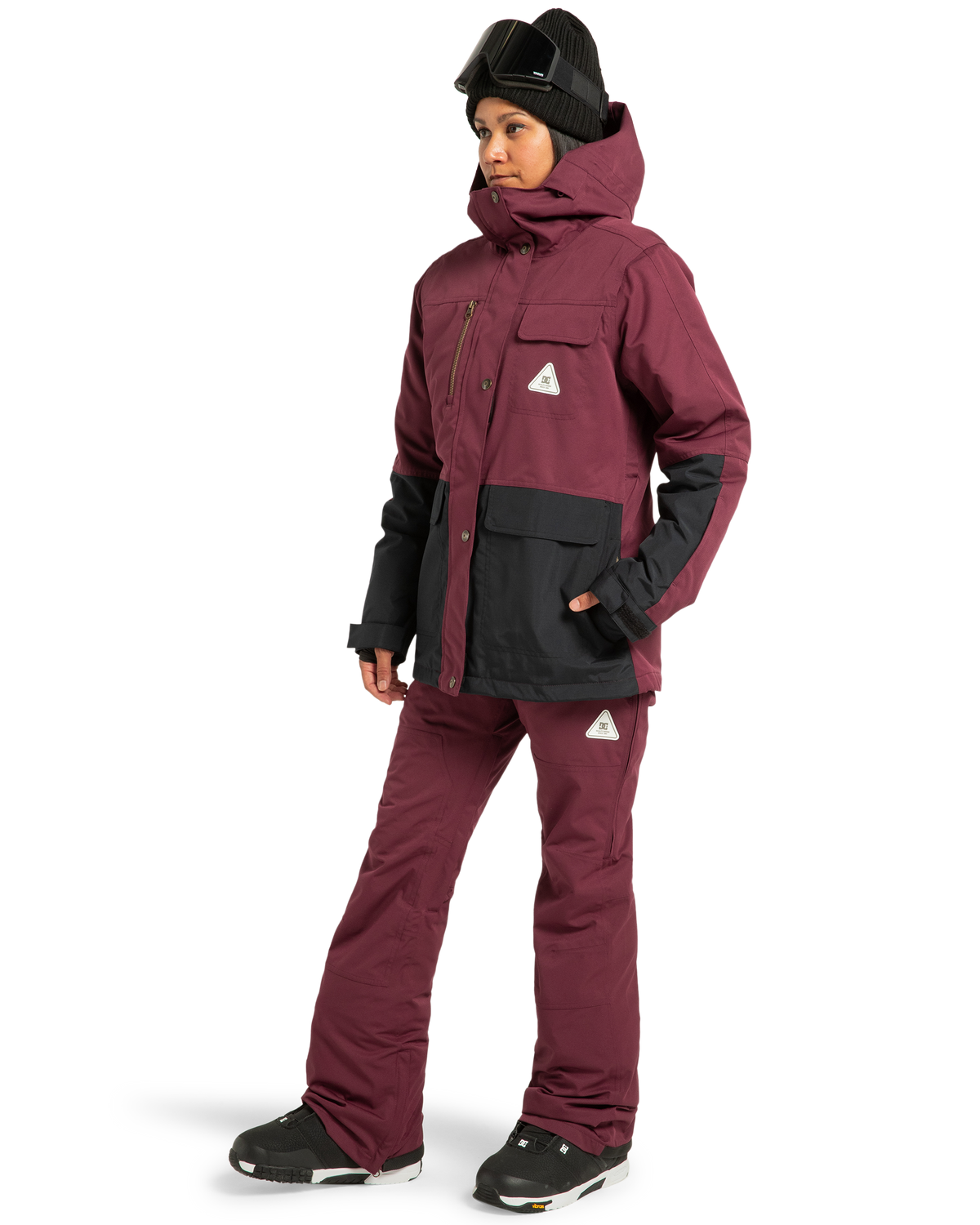 A woman wearing the DC Shoes Women’s Crusade Shell Bib in maroon and black, paired with a matching jacket, black gloves, black winter boots, and a black beanie with ski goggles, stands against a white background.