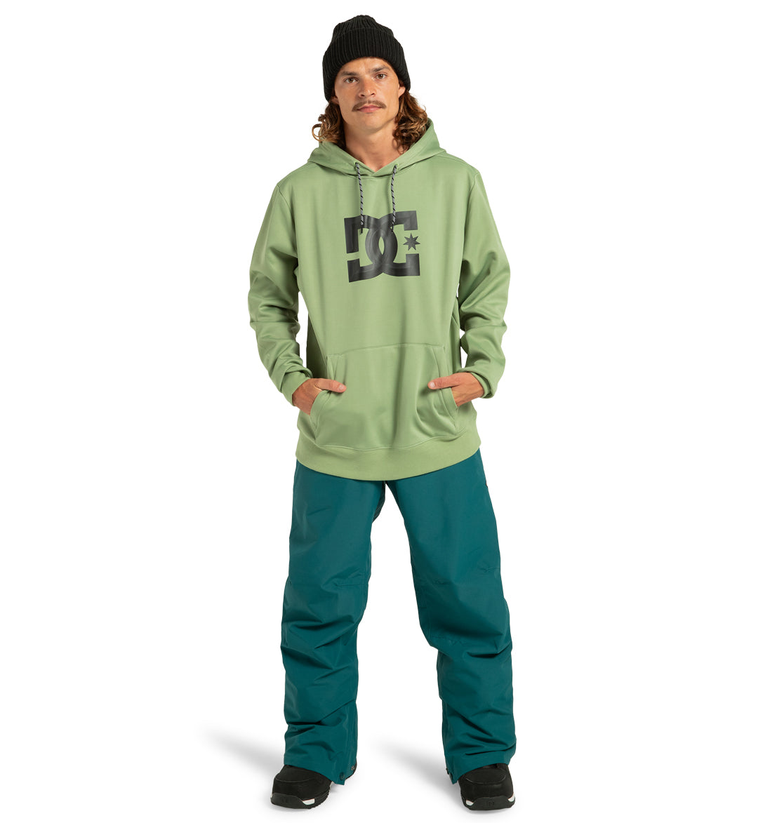 A person stands facing forward against a white background, hands in the pocket of a light green DC Shoes Unisex Snowstar Hoodie—a durable polyester hoodie designed with weather defense—paired with a black beanie, teal snow pants, and black shoes.