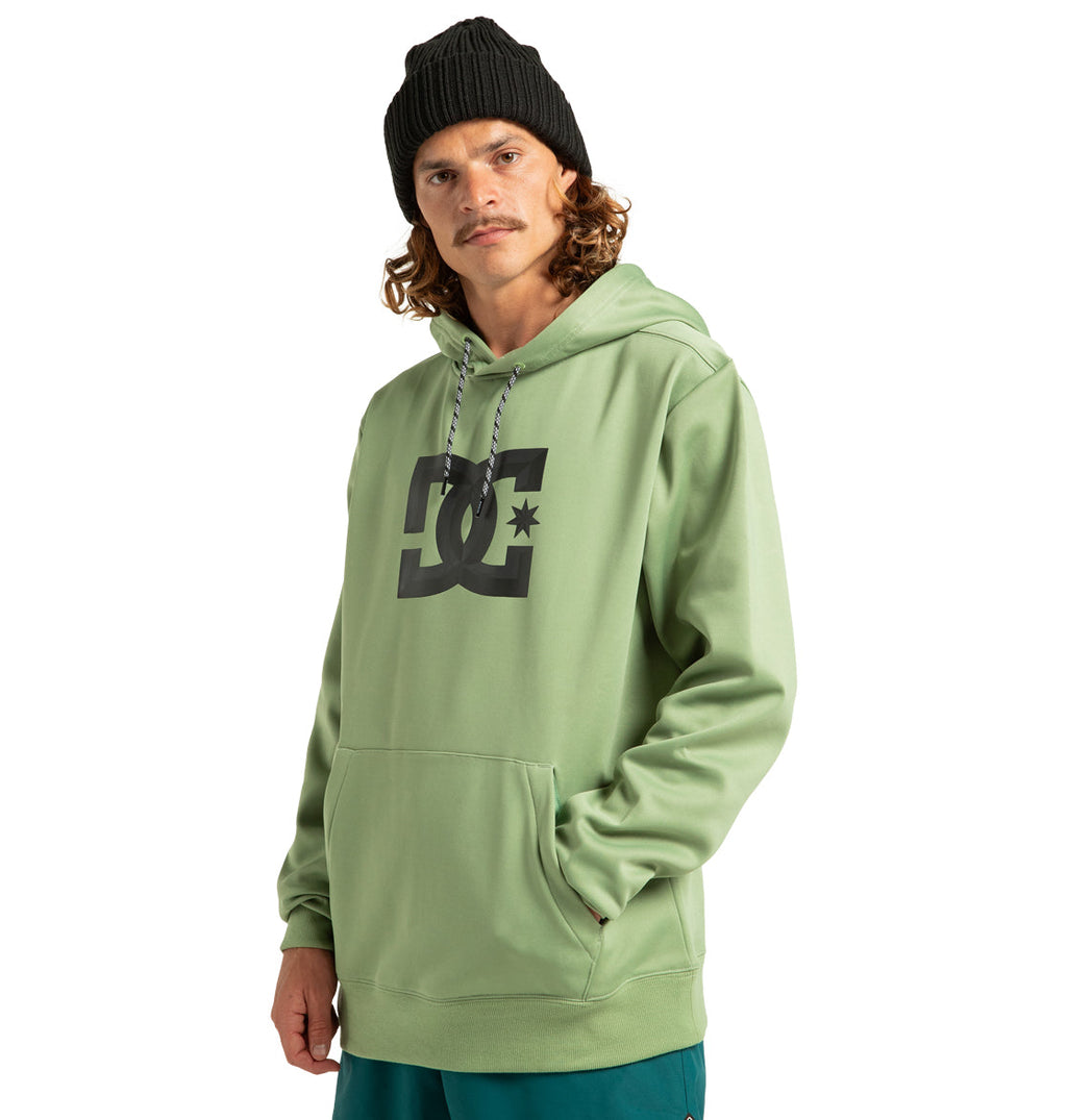 A man stands facing forward against a plain white background, wearing a light green DC Shoes Unisex Snowstar Hoodie made from weather defense polyester, paired with dark green pants and a black beanie, with one hand in his pocket.