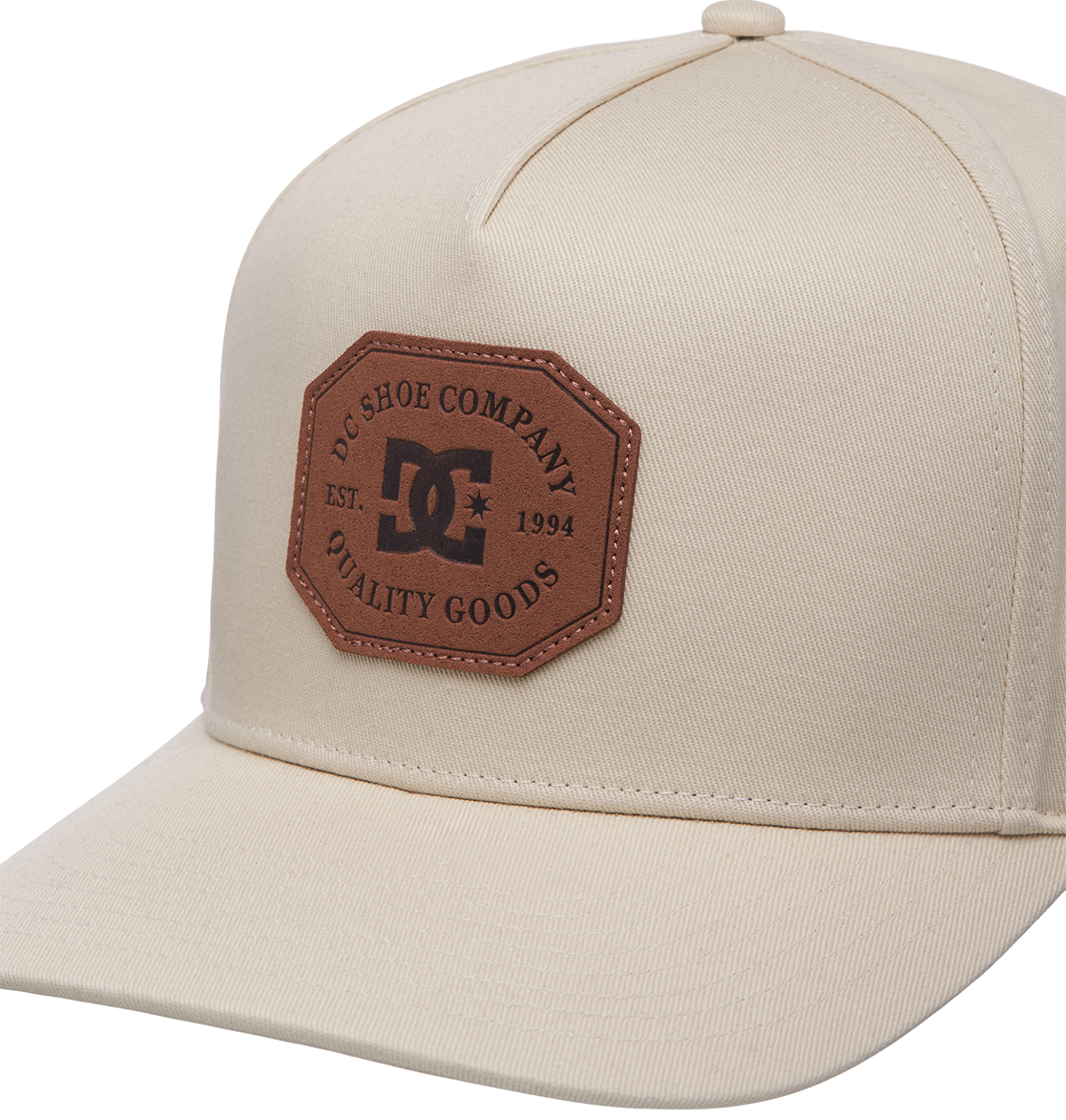 The Men's Reynotts Cap by DC Shoes is a beige skate cap featuring a brown hexagonal patch on the front with DC branding, reading "DC Shoe Company Quality Goods Est. 1994," and has a classic curved brim.