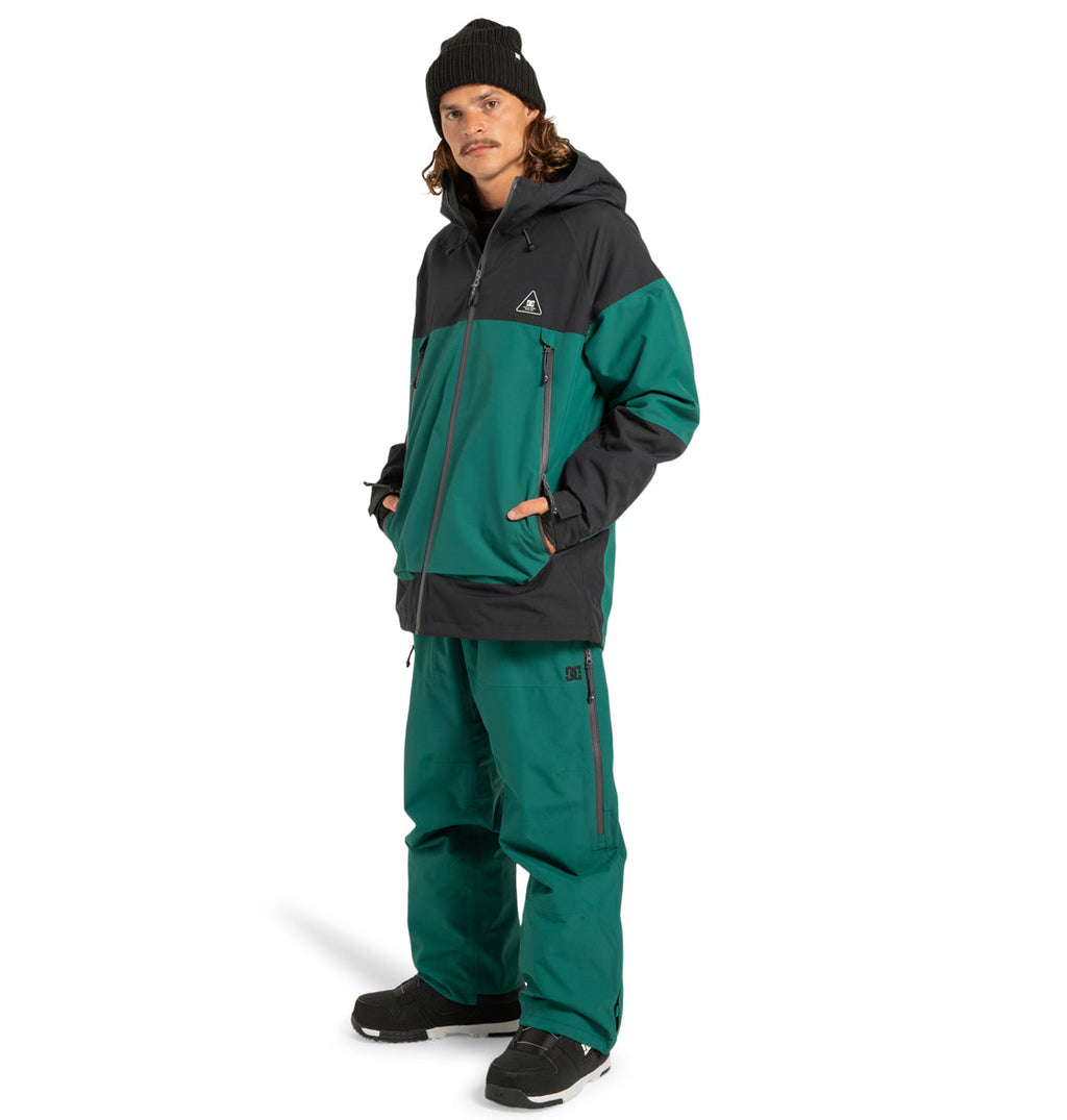 A person wearing a black beanie, black shoes, teal pants, and the DC Shoes Unisex Intel 30K Insulated Jacket in a teal and black colorway stands with hands in pockets against a white background—perfectly outfitted for adventure with built-in weather defense.