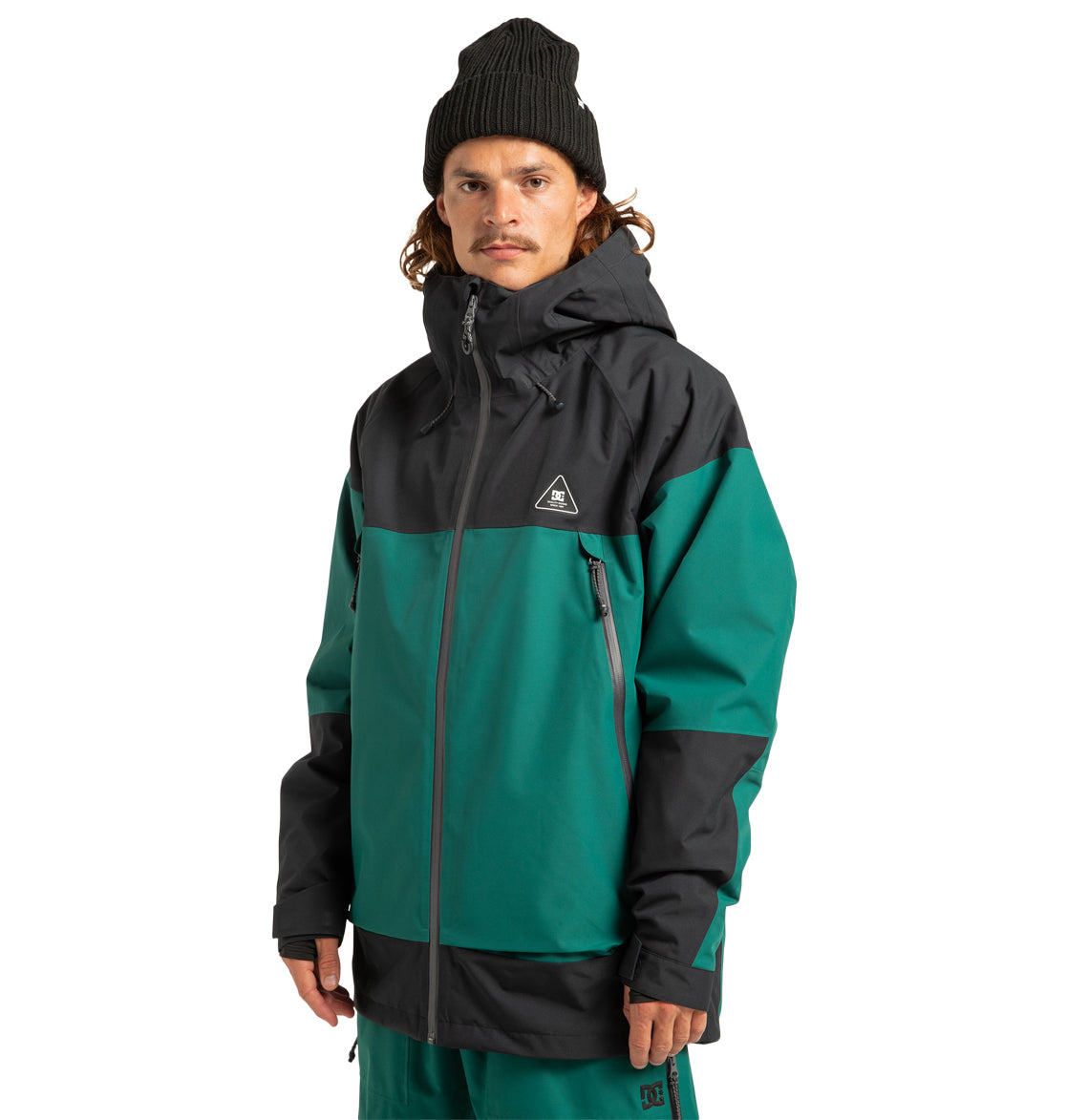 A person with long hair and a mustache wears a black beanie and the DC Shoes Unisex Intel 30K Insulated Jacket in green and black—perfect for weather defense—standing against a plain white background.