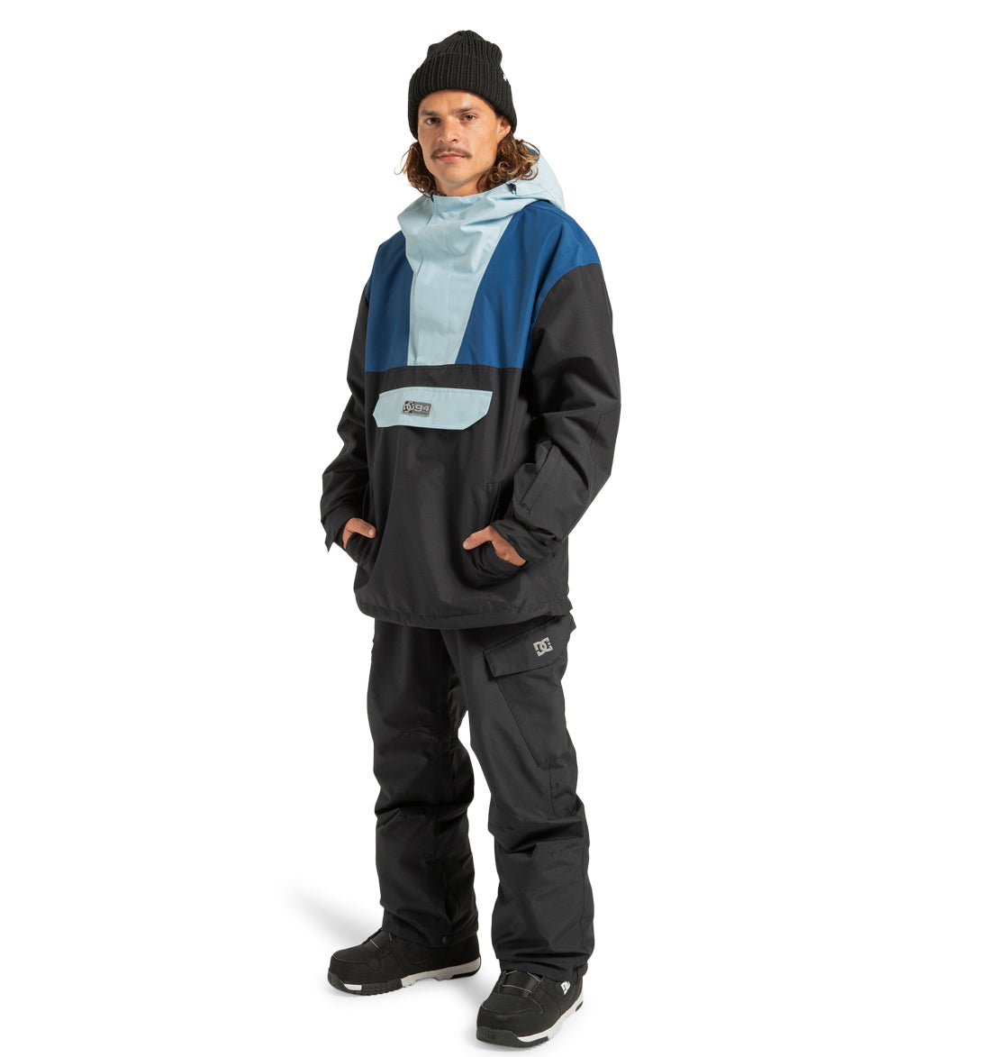A person wearing a black beanie, black snow pants, and black boots stands facing forward with hands in pockets against a white background, showcasing the Men’s DC-43 Anorak Jacket by DC Shoes—a blue and black color-blocked outdoor adventure jacket.