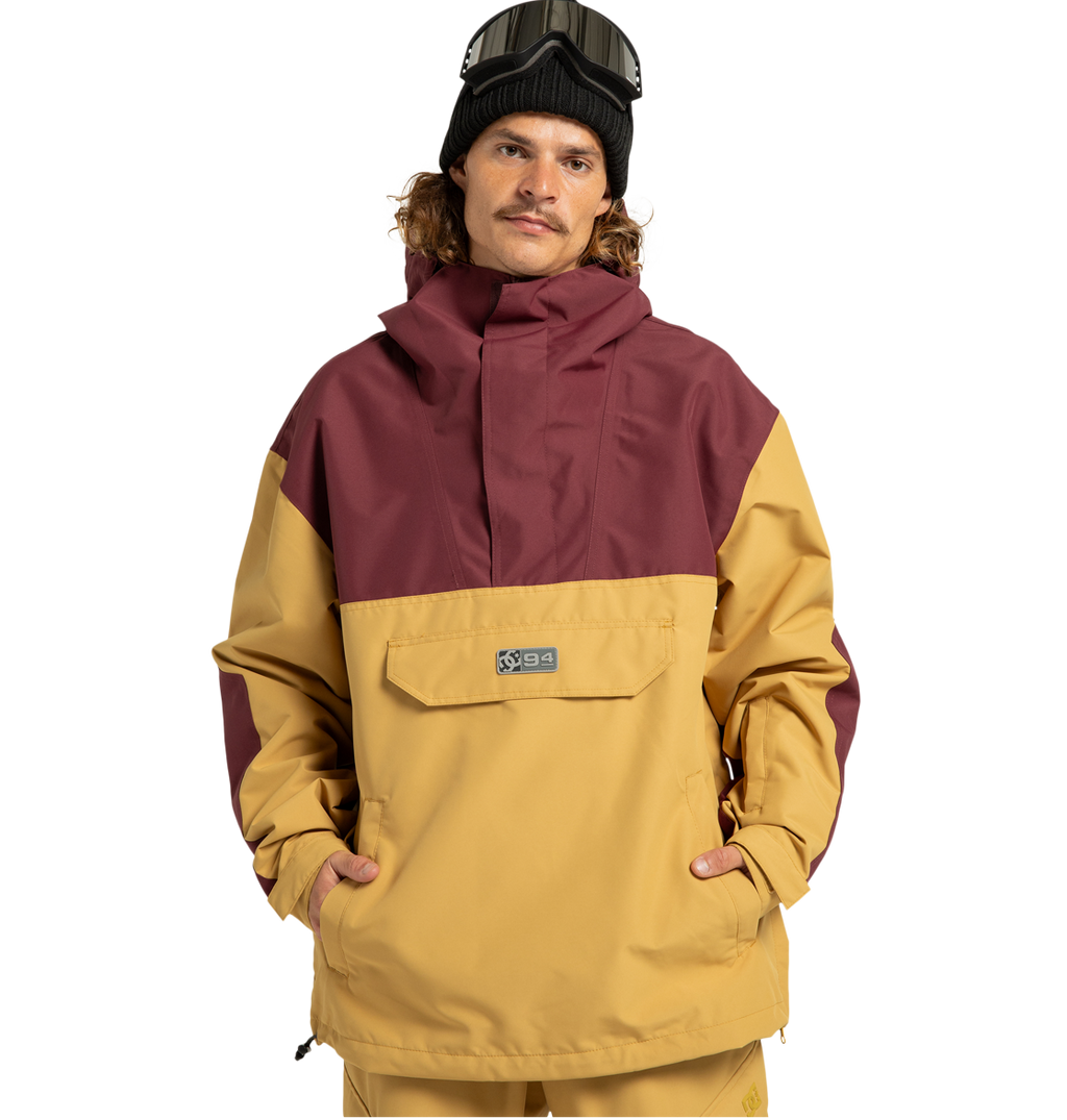 A person wearing a mustard yellow and maroon Men’s DC-43 Anorak Shell Jacket by DC Shoes, featuring a front pocket, tan pants, a black beanie, and ski goggles on their head stands with hands in pockets against a plain white background. This stylish jacket is made from recycled polyester and functions as a versatile weather defense shell, providing comfort and protection in any condition.