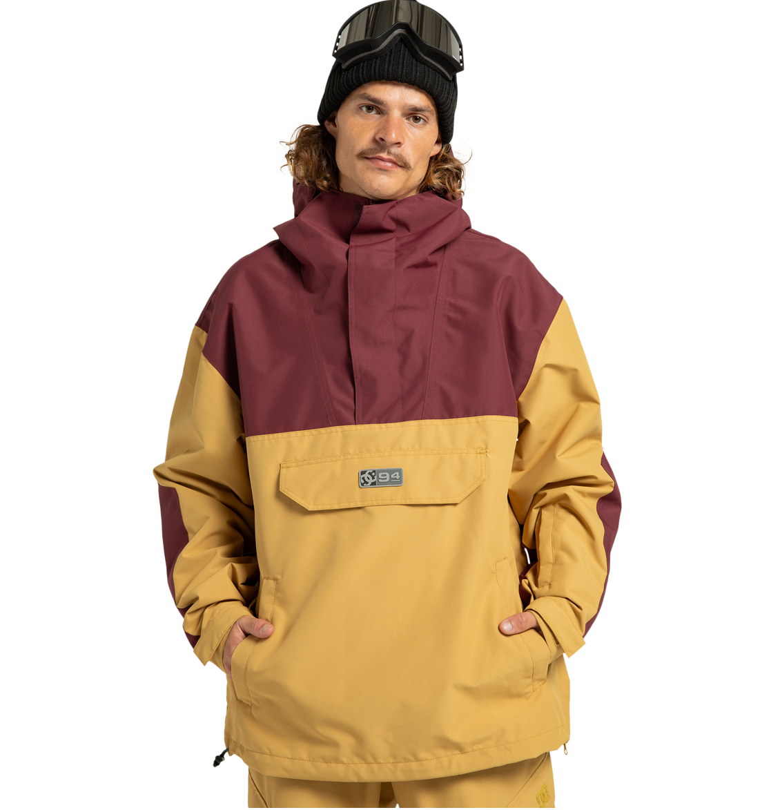 A person wearing a mustard yellow and maroon Men’s DC-43 Anorak Shell Jacket by DC Shoes, featuring a front pocket, tan pants, a black beanie, and ski goggles on their head stands with hands in pockets against a plain white background. This stylish jacket is made from recycled polyester and functions as a versatile weather defense shell, providing comfort and protection in any condition.