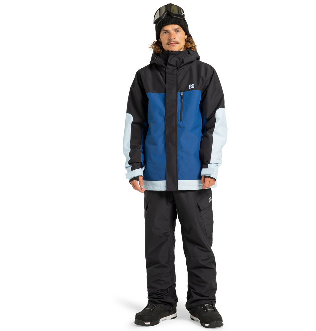 A person wearing the DC Shoes Men’s Defiant Insulated Jacket—featuring a colorblock design in blue, black, and light blue with recycled polyester construction and Weather Defense 10 technology—is paired with black snow pants, black boots, a black beanie, and ski goggles while standing against a plain white background.