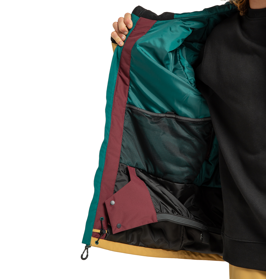 A person holds open the DC Shoes Men’s Defiant Insulated Jacket, revealing its teal, burgundy, and yellow interior with a mesh pocket and snap buttons. The person is wearing a black sweatshirt underneath.