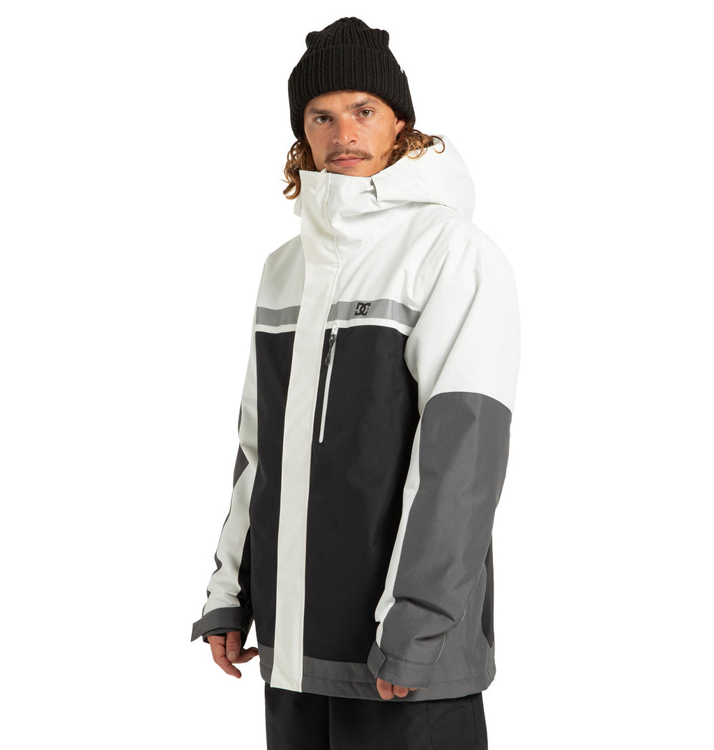 A man wearing a black beanie and the Men’s Defiant Insulated Jacket by DC Shoes—featuring a color-blocked design in white, black, and gray, crafted from recycled polyester—stands facing slightly left, looking at the camera with a neutral expression, ready for his next winter adventure.