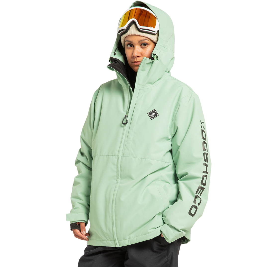 A person is wearing the DC Shoes Unisex Basis Insulated Jacket in light green, with the hood up, paired with black pants and ski goggles resting on their head. “DC SHOES” is printed on the left sleeve. Crafted from recycled polyester, this jacket is designed for demanding winter pursuits. The background is plain white.