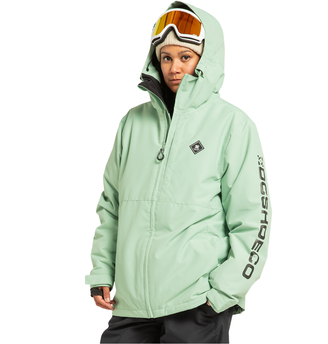 A person is wearing the DC Shoes Unisex Basis Insulated Jacket in light green, with the hood up, paired with black pants and ski goggles resting on their head. “DC SHOES” is printed on the left sleeve. Crafted from recycled polyester, this jacket is designed for demanding winter pursuits. The background is plain white.