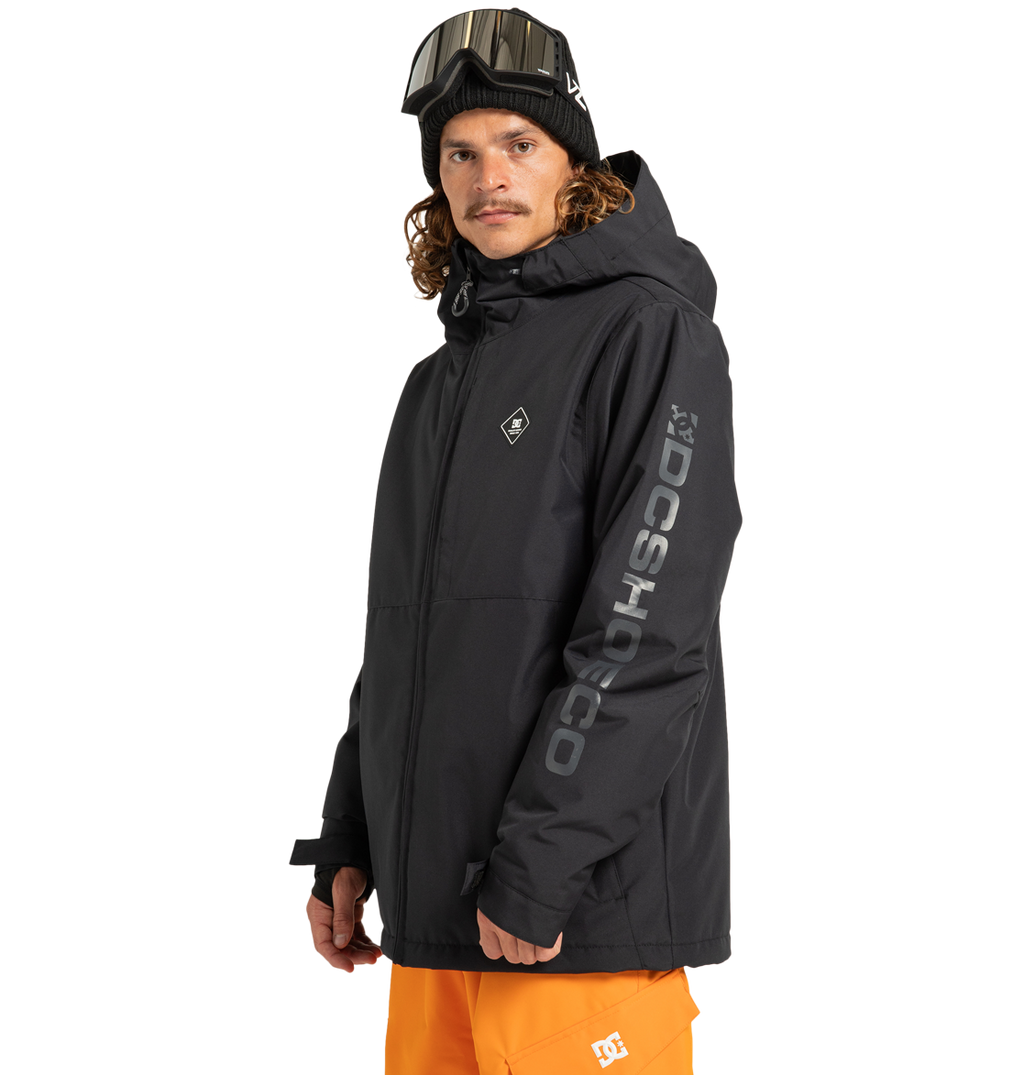 A person wearing the Unisex Basis Insulated Jacket by DC Shoes—a black, insulated outdoor jacket crafted from recycled polyester with “DCSHOECO” branding on the sleeve—paired with orange pants, ski goggles on their head, and a black beanie, stands against a plain white background.