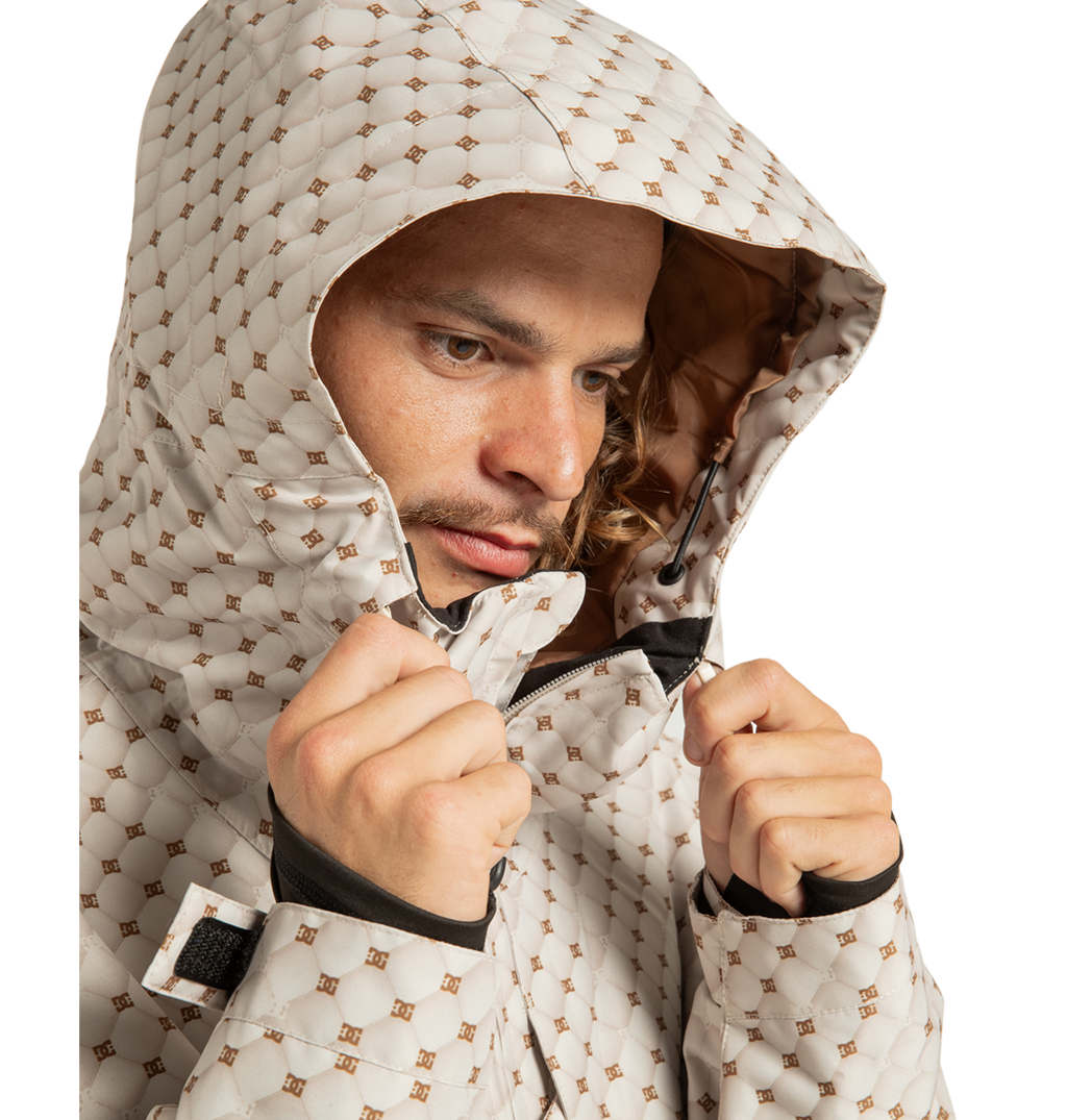 A person wearing the DC Shoes Unisex Basis Print Insulated Jacket, made from recycled polyester, zips up the light-colored geometric print hooded jacket while looking down and slightly to the side, with the hood pulled over their head.