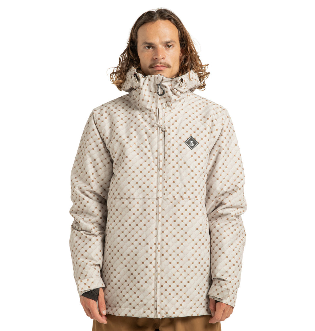 A person with long curly hair is wearing the DC Shoes Unisex Basis Print Insulated Jacket—an insulated beige jacket with a diamond pattern, hood, chest logo, and zip front made from recycled polyester—standing facing forward against a plain white background.