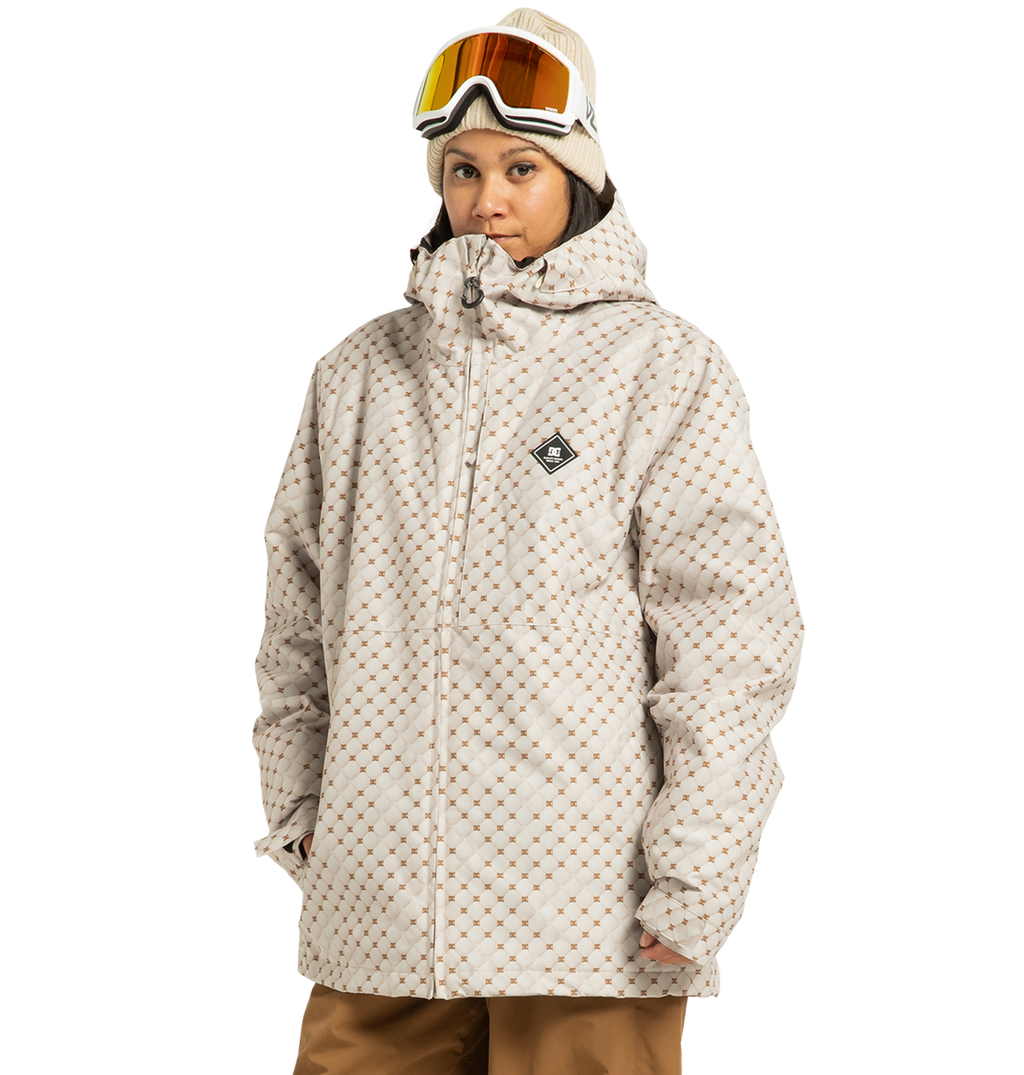 A person stands against a white background wearing the DC Shoes Unisex Basis Print Insulated Jacket—crafted from recycled polyester with a beige patterned design—paired with tan pants, a beige beanie, and ski goggles.