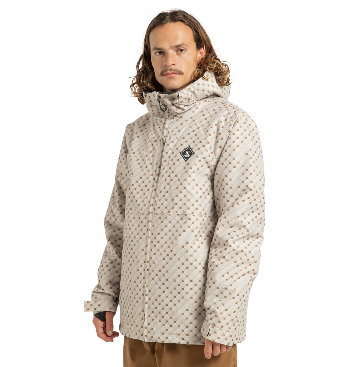 A man with long curly hair is wearing the DC Shoes Unisex Basis Print Insulated Jacket, made from recycled polyester in a light beige patterned design, paired with tan pants, standing against a plain white background.
