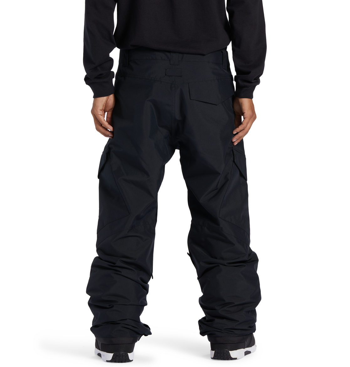 A man stands facing away, wearing DC Shoes Men’s Banshee Insulated Pants—loose-fitting black cargo pants designed for winter, featuring large pockets and covering the tops of his shoes—and a black long-sleeve shirt, with his hands relaxed at his sides.
