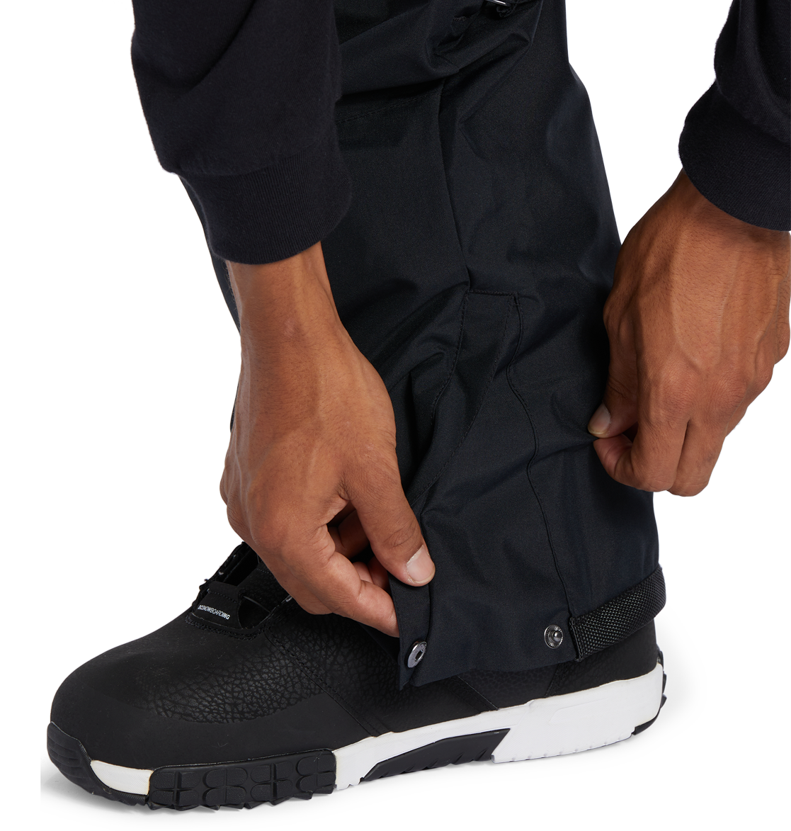 A person adjusts the snap button on the cuff of DC Shoes Men’s Banshee Insulated Pants, worn over black and white outdoor boots—an essential piece of winter gear.