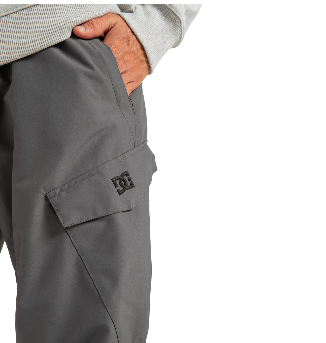 A person wears the Men’s Banshee Insulated Pants by DC Shoes—gray pants featuring a flap pocket with the DC logo, made from durable recycled polyester. Their left hand rests in the pocket, pairing the ideal men’s insulated winter mountain wear with a light gray long-sleeve shirt.