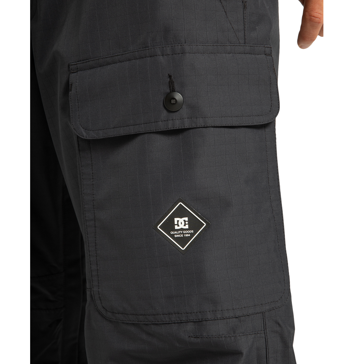 Close-up of a black cargo pocket on the DC Shoes Men’s Code Shell Pants, featuring a buttoned flap and a diamond-shaped patch with the DC logo and “Quality Goods” on the pocket.