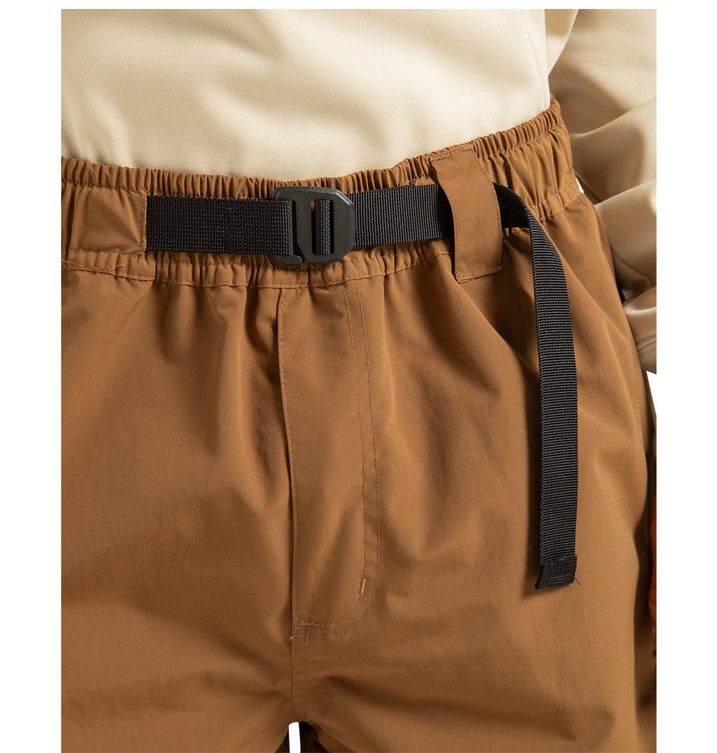 A close-up shows a person wearing tan DC Shoes Unisex Primo Shell Pants, which feature an elastic waistband and a black adjustable belt with a plastic buckle. Designed for outdoor enthusiasts, these pants offer weather defense properties. The person pairs them with a beige top.