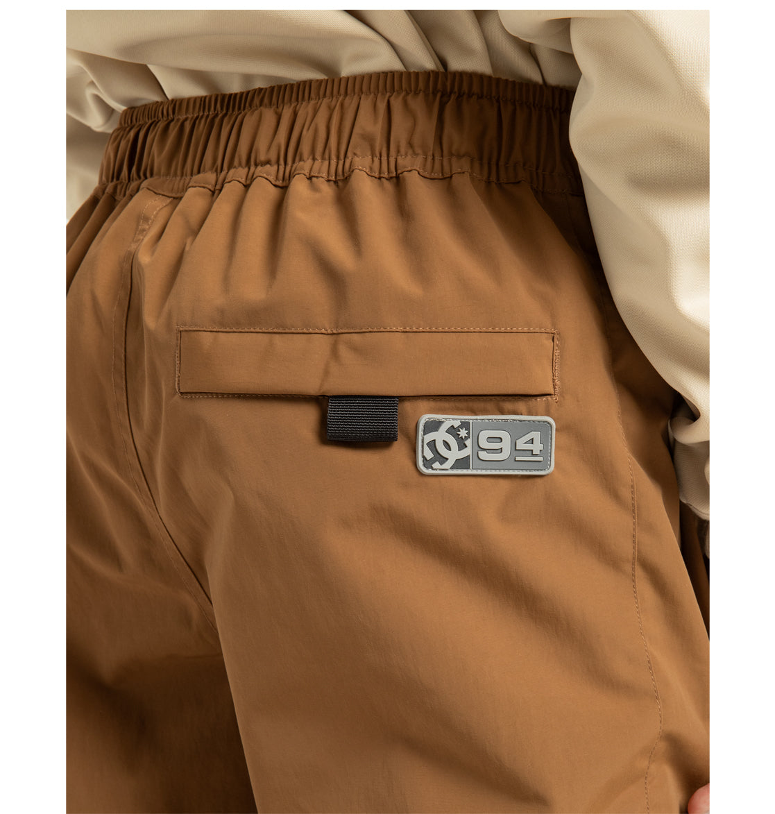 Close-up of a person wearing light brown Unisex Primo Shell Pants from DC Shoes, featuring an elastic waistband, back pocket, and a ‘94’ patch with a gear symbol on the right side—engineered for weather defense to meet the needs of outdoor enthusiasts. The person is also wearing a cream-colored top.