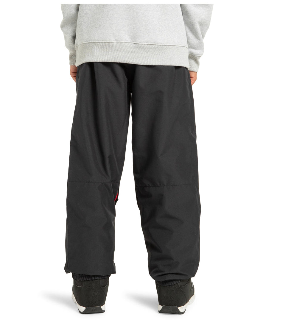 A person wearing DC Shoes’ Unisex Primo Snow Pants in black, paired with a grey sweatshirt and black-and-white sneakers, stands with their back to the camera. Only the lower half of their body is visible.