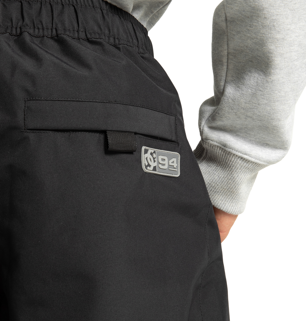 A close-up of a person wearing DC Shoes Unisex Primo Snow Pants in black, featuring a patch with a graphic and the number 94. The person’s hand is by their side, and they are also wearing a light gray sweatshirt.