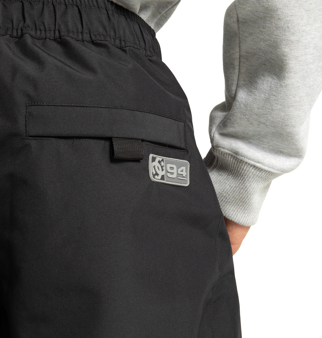A close-up of a person wearing DC Shoes Unisex Primo Snow Pants in black, featuring a patch with a graphic and the number 94. The person’s hand is by their side, and they are also wearing a light gray sweatshirt.