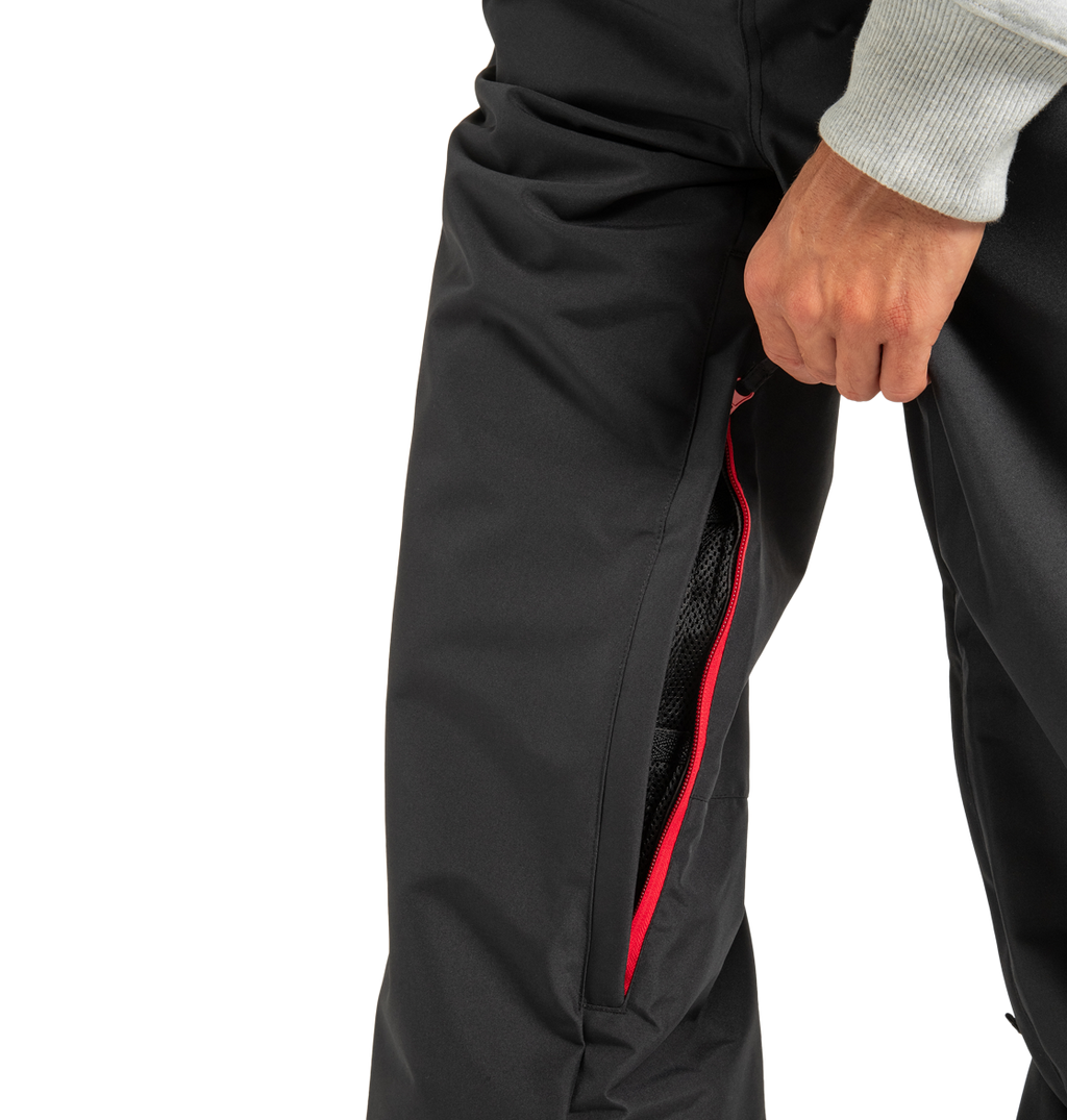 A person wearing DC Shoes Unisex Primo Snow Pants is shown with the red-zippered ventilation partially unzipped, revealing the mesh lining inside, as they hold the zipper with one hand. Only their lower torso and hand are visible.