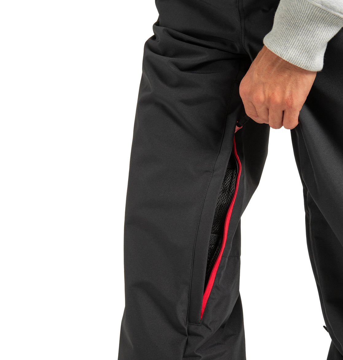 A person wearing DC Shoes Unisex Primo Snow Pants is shown with the red-zippered ventilation partially unzipped, revealing the mesh lining inside, as they hold the zipper with one hand. Only their lower torso and hand are visible.