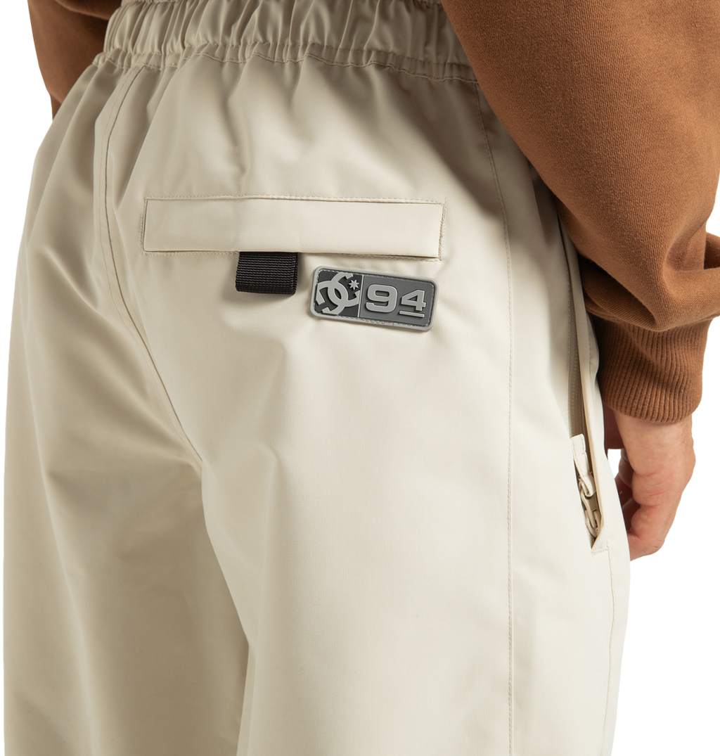 A close-up of a person wearing the DC Shoes Unisex Primo Snow Pants in beige, featuring an elastic waistband, a back pocket, and a patch reading “94” beside a circular logo. Made from recycled polyester, these waterproof snow pants are styled with a brown top.