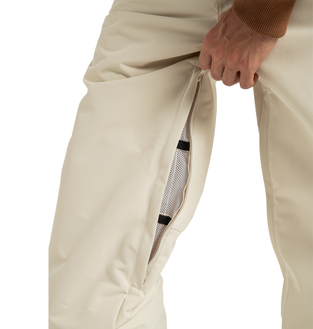 A person unzips a vent on the side of cream-colored DC Shoes Unisex Primo Snow Pants, revealing a mesh lining made from recycled polyester beneath. Only the lower torso and hand are visible.